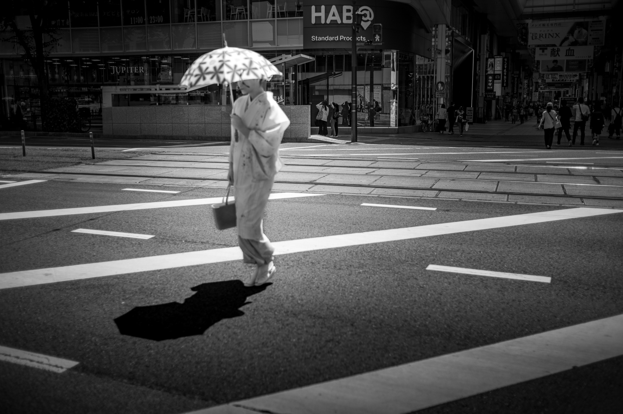 A figure in kimono crosses an empty street under a parasol, the shadow stretched long on the asphalt.
